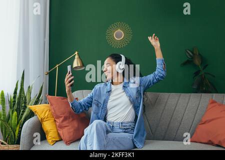 Attraente giovane donna ispanica che ascolta musica in cuffie bianche. Si siede sul divano, tiene il telefono nelle mani, prende un selfie, prende un video, alza le mani, balla. Foto Stock