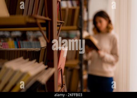 Ragazza offuscata del college che legge un libro mentre si trova accanto alle librerie nella biblioteca Foto Stock