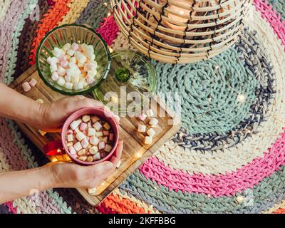 Vista ravvicinata delle mani delle donne che tengono una tazza rossa di cacao con mini marshmallows in un'accogliente giornata invernale, lanterna e luci a LED accese. Copia spazio su h Foto Stock