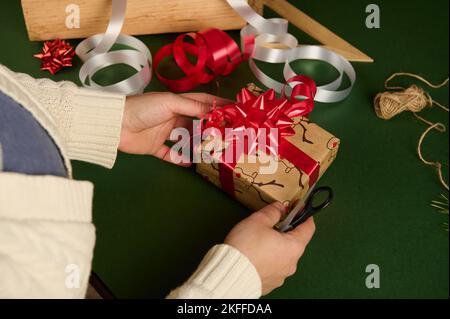 Vista dall'alto delle mani della donna che legano l'arco bello da un nastro rosso lucido mentre decorano un regalo di Natale. Avvolgimento. Foto Stock