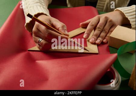 Le mani della donna con il pastello di legno e il righello triangolare disegnano sulla carta da imballaggio, calcolano la quantità di esso per i regali di imballaggio Foto Stock