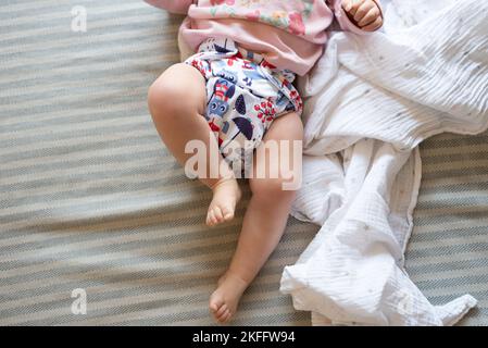 Un bambino sdraiato su un letto con la messa a fuoco sulle gambe del bambino e pannolino in tessuto riutilizzabile con una bella stampa Foto Stock