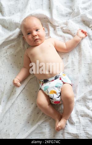 Una ragazza felice e soddisfatta del bambino che giace sulla sua parte posteriore. Indossa un pannolino in tessuto moderno e riutilizzabile con una bella stampa Foto Stock