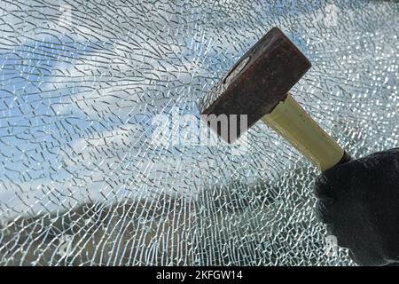 Mani guazzate di un criminale distruggendo una finestra di vetro laminato con un martello, violenza, furto e vandalismo concetto, copia spazio, selezionato fuoco Foto Stock