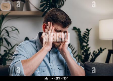 Uomo bearded sovraccarico con mal di testa seduto sul divano in camera. Triste stanco operaio caucasico maschio sensazione sfinito dal lavoro eccessivo. Ragazzo che lavora in remoto f Foto Stock