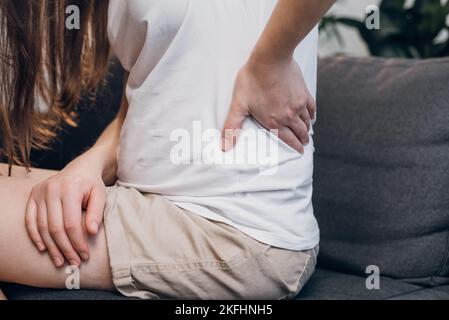 Primo piano di una giovane donna malsana che soffre di mal di schiena, osteocondrosi toccando la schiena con la mano seduta sul divano grigio a casa Foto Stock