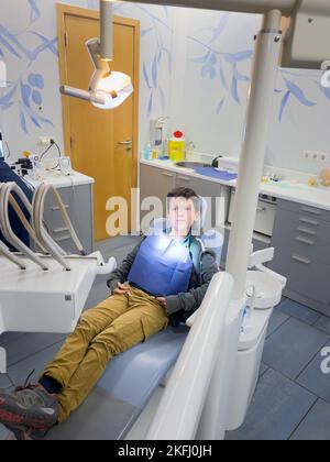 Ragazzo caucasico che indossa il bavaglino blu che guarda lontano mentre aspetta il medico sulla sedia del dentista in clinica dentale Foto Stock