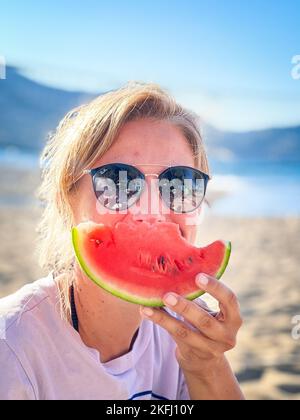 Primo piano ritratto di una donna media adulta che indossa occhiali da sole e mangia una fetta di anguria in spiaggia durante le vacanze estive Foto Stock