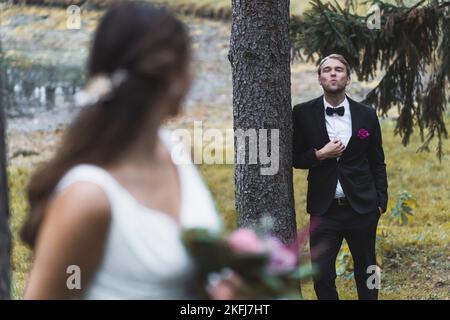 Giovane adulto scandinavo in abito elegante che soffia un bacio alla sua nuova bella moglie turca mentre si appoggia su un albero. Sposa sfocata in primo piano. Concentratevi sullo spazio per lo sposo sullo sfondo. Foto di alta qualità Foto Stock