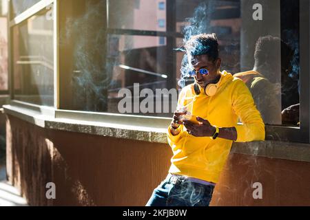 Un giovane uomo africano vestito con una felpa gialla, jeans blu e occhiali da sole si appoggia su una sporgenza della finestra guardando il suo telefono delle cellule mentre tiene una c Foto Stock