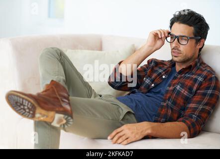 Hippster appendere fuori. Un bellissimo giovane uomo alla moda sdraiato su un divano a casa con le gambe incrociate indossando occhiali. Foto Stock