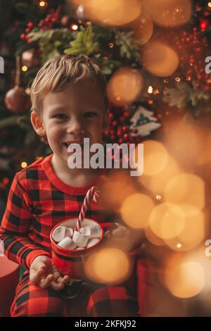 Felice bambino sognatore in attesa di Miracle Babbo Natale godere dolci colore lecca. Rosso a scacchi Sleepwear ragazzo allegro celebrare nuovo anno Hold Natale Cup Foto Stock