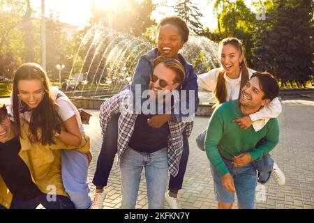 Amici gioiosi che si divertono, ridono e si trasportano a vicenda sul viale della città in una giornata calda. Foto Stock