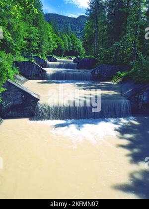 Affluente fluviale fangoso dalle vette delle Alpi dolomitiche dopo forti piogge. Il letto del fiume alimenta la corrente alla centrale idroelettrica e su Foto Stock