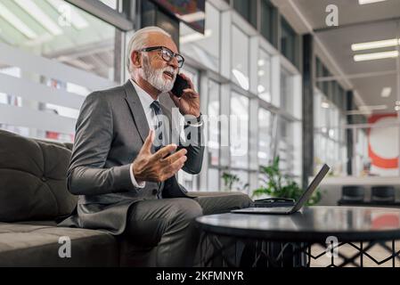 Sorridente professionista di sesso maschile gesturing mentre si parla su smartphone. L'imprenditore senior utilizza il notebook mentre è seduto al tavolo. Sta indossando il vestito mentre Foto Stock