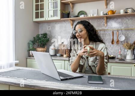 Giovane bella donna d'affari latino-americana, freelance che lavora a casa on-line in cucina con un computer portatile. Comunica tramite videochiamata. Contiene una tazza di caffè. Foto Stock