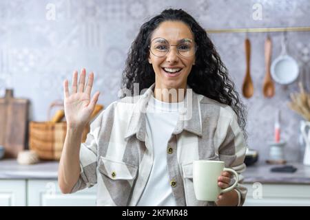 Giovane bella donna ispanica che parla e agita alla telecamera durante una videochiamata. In piedi in cucina a casa, tenendo una tazza, sorridendo, gestisce il suo blog. Foto Stock