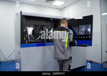 Stampante industriale a polvere 3D per metallo e operatore. Uomo in tuta protettiva vicino alla finestra di una grande stampante 3D in laboratorio. Le mani indossano grandi maniche nere per sigillare le lavorazioni all'interno della macchina Foto Stock