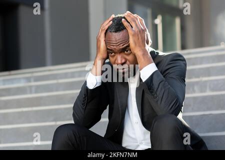 Rovesciato e licenziato lavoratore di ufficio seduto sulle scale fuori ufficio edificio primo piano uomo d'affari in costume depresso perso denaro fallita afro-americano lavoratore seduto triste. Foto Stock