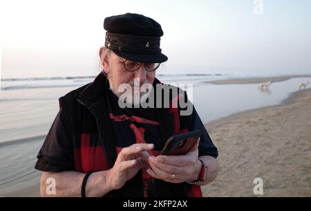 Un uomo anziano con il cappello dei capitani, gli occhiali e la barba è occupato con il suo smartphone. È in spiaggia. I cani giocano in background Foto Stock