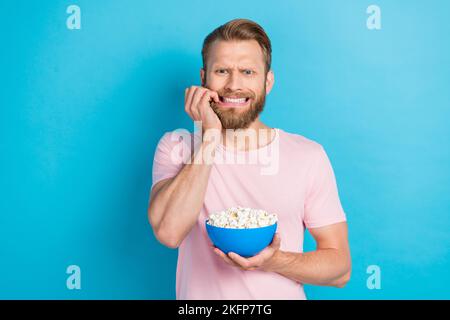 Foto di divertente uomo spaventato indossare t-shirt rosa mordente dita guardare orrore mangiare pop mais isolato sfondo di colore blu Foto Stock