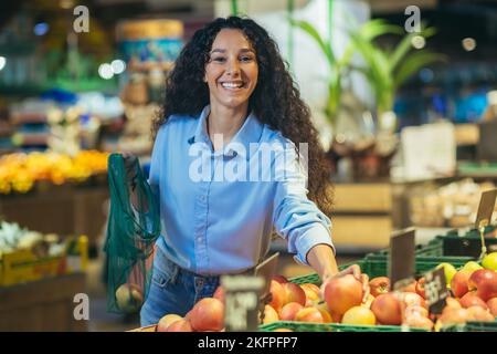 Ritratto di donna felice acquirente in supermercato, donna ispanica sceglie mele e frutta sorridendo e guardando la macchina fotografica, con il cestino della drogheria sceglie le merci. Foto Stock