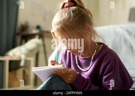 Bambina che scrive i suoi segreti nel suo diario mentre si siede nella camera da letto Foto Stock