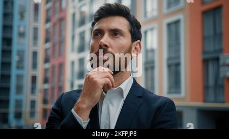 Caucasian intelligente perplesso uomo premuroso tenere mano al mento sognando sulla crescita di carriera datore di lavoro di successo in elegante vestito nero stand fuori moderno Foto Stock