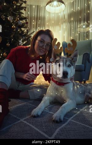 Giovane donna la sera di Natale insieme con il cane da compagnia che ha divertimento mettere su corna di cervo di Natale e ghirlande seduti sul pavimento al Natale Foto Stock