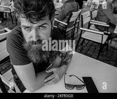 Un ragazzo che si riposa sulla terrazza del caffè. Hipster, cliente, visitatore sul serio si siede al caffè all'aperto. Uomo con barba e baffi di umore calmo Foto Stock