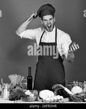 Cuoco lavora in cucina vicino a verdure e utensili. Uomo in cappello di  cuoco e grembiule trita pepe. Cucina e cucina. Chef con viso attento tiene  il telefono sulla spalla e coltello