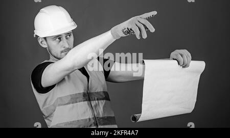 Uomo in casco, elmetto tiene la pianta di costruzione, comandi lavori, sfondo rosso. Concetto di caposquadra. Ingegnere, architetto, costruttore su faccia rigida tiene vecchio Foto Stock