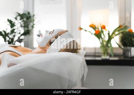 Vista laterale interna ritratto di positiva donna caucasica rilassata nel suo 30s sdraiato sul letto SPA e godendo il suo giorno SPA. Piante e tulipani sfocati sullo sfondo. Foto di alta qualità Foto Stock