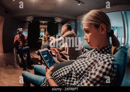 Donna incinta seduta sul divano di un moderno ufficio circondato da colleghi e utilizzando un tablet Foto Stock