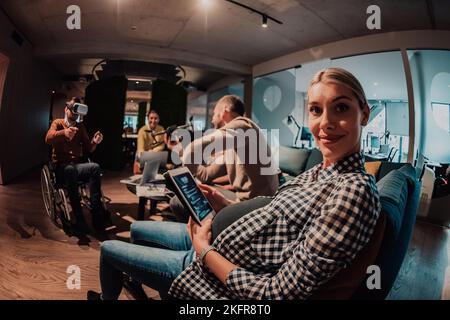 Donna incinta seduta sul divano di un moderno ufficio circondato da colleghi e utilizzando un tablet Foto Stock