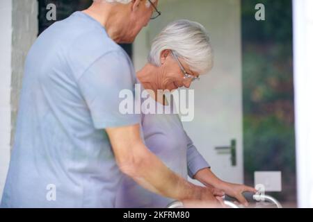 Un uomo anziano che aiuta la moglie con il suo camminatore ortopedico. Foto Stock