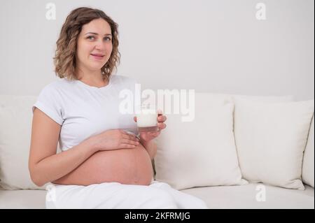 Una donna incinta siede su un divano bianco e beve latte. Foto Stock