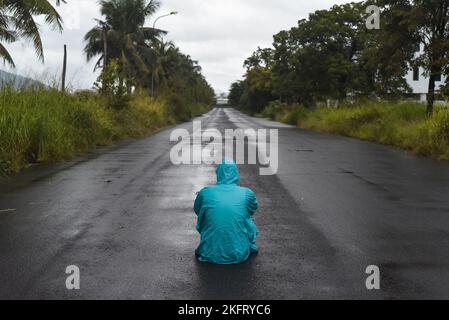 Concetto depressivo con l'uomo solitario in impermeabile seduto su strada bagnata sotto la pioggia Foto Stock