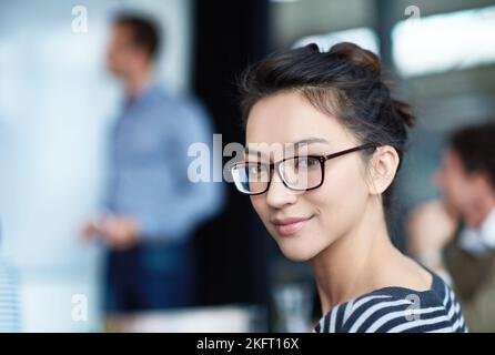 La sua traiettoria di carriera non conosce limiti. Ritratto di una giovane donna attraente seduta in un ufficio con i colleghi sullo sfondo. Foto Stock