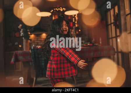 Attiva ragazza bambino giappone con elegante modello di capelli brunette lunga posa a casa in bokeh ghirlands noel. Asian piccolo fashionista in rosso plaid pajama danza Foto Stock