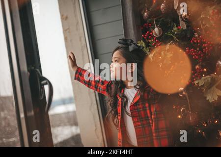 Felice giappone piccola ragazza sognatore guardando attraverso la finestra e miracolo in attesa da Santa posa vicino a noel albero. Bambino asiatico in abito rosso plaid festeggiamento Foto Stock