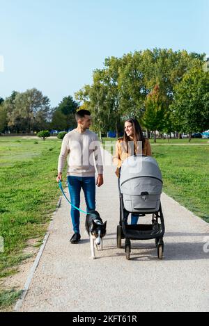 Felice coppia con passeggino e cane che camminano insieme sul sentiero contro prato verde e alberi durante la passeggiata in estate Foto Stock