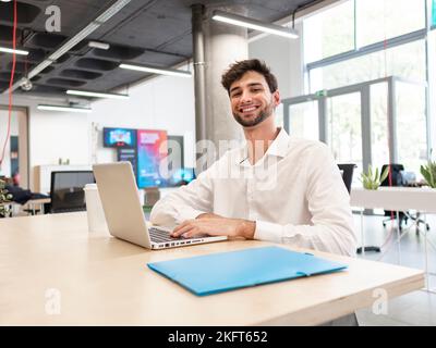 Giovane uomo bearded concentrato in camicia bianca seduto al tavolo e navigando in internet via computer portatile mentre si lavora su un computer portatile in una spaziosa caffetteria ufficio Foto Stock