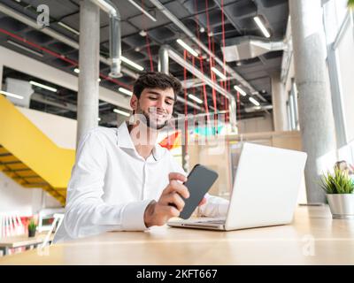 Giovane uomo d'affari concentrato con beared in camicia bianca seduto al tavolo con smartphone e digitando le informazioni sul laptop in un'area di lavoro spaziosa e luminosa Foto Stock