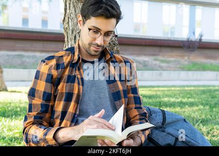 Studente maschile ispanico concentrato in camicia a scacchi seduta su erba verde vicino allo zaino e la lettura interessante libro mentre si appoggia sul tronco dell'albero Foto Stock