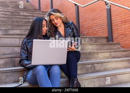 Studenti di sesso femminile multietnica con computer portatile seduto su passi e discutere i dati in blocco note mentre fare i compiti insieme sulla strada Foto Stock