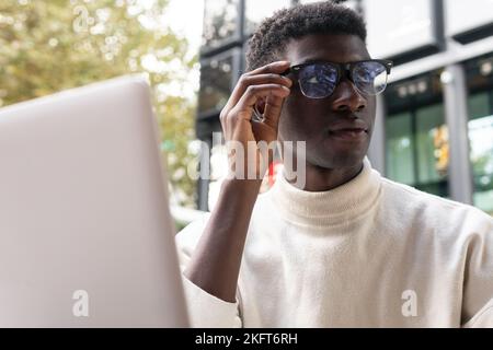 Libero professionista afro-americano concentrato in occhiali seduti al tavolo con computer portatile al caffè all'aperto Foto Stock