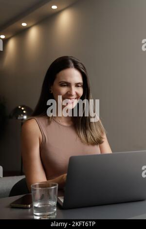 Fidata freelancer femminile di mezza età con lunghi capelli castani in un abbigliamento casual sorridente mentre scrivi sul computer portatile durante il lavoro online a casa Foto Stock