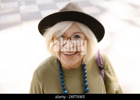 Ritratto di donna anziana sorridente in occhiali e perle in piedi sulla strada vicino a casa e guardando la macchina fotografica Foto Stock