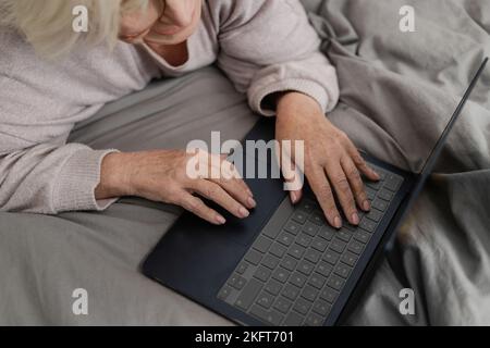 Da sopra anonima anziana signora sdraiata su un morbido letto con lenzuola grigie e informazioni di digitazione sul computer portatile a casa Foto Stock